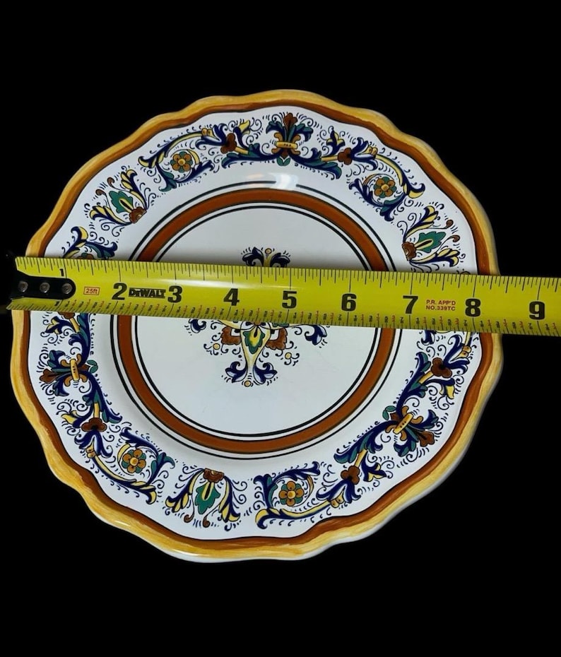 May include: A decorative ceramic plate with a scalloped edge. The plate features a white center with a brown border and intricate floral designs in blue, green, and yellow. The plate measures approximately 8 inches in diameter.