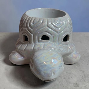 Może przedstawiać: Iridescentny ceramiczny świecznik w kształcie żółwia. Ozdoba ma perłowe wykończenie, z dużym otworem na świecę i wyciętymi otworami na oczy. Żółw ma cztery nogi i szczegółowy pancerz.