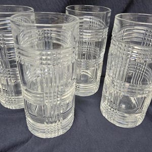 May include: Four clear, patterned drinking glasses. Each glass has a cylindrical shape with a textured, grid-like design. The glasses are made of transparent glass and are arranged on a dark blue surface.