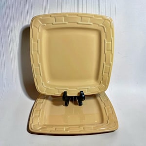 May include: Two square, pale yellow ceramic plates featuring a raised, embossed basket-weave pattern along the wide, flat rims. One plate is positioned upright on a black plastic stand, while the second plate rests horizontally beneath it.