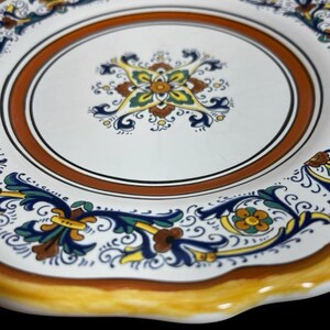 May include: White ceramic plate with a decorative floral design. The plate features a brown and orange border, with blue, yellow, and green floral patterns. The rim is scalloped and painted yellow.