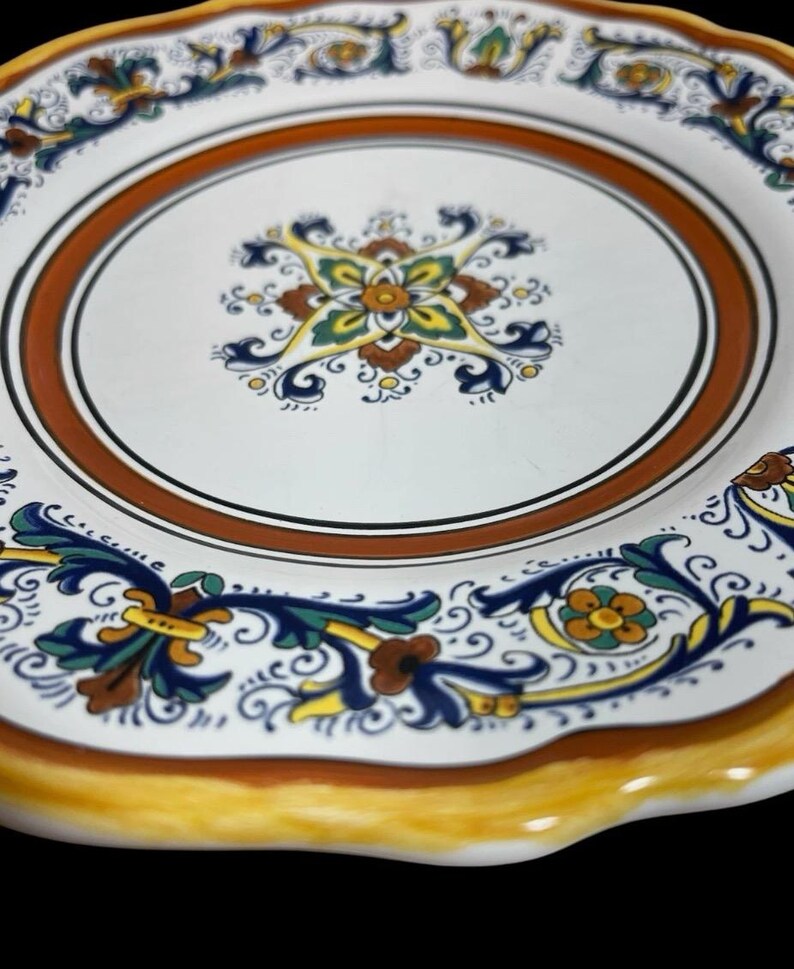 May include: White ceramic plate with a decorative floral design. The plate features a brown and orange border, with blue, yellow, and green floral patterns. The rim is scalloped and painted yellow.
