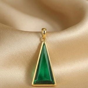 以下が含まれることがあります： 三角形のエメラルドグリーンの宝石が付いた、金色のペンダント。ペンダントはシンプルでエレガントなデザインで、グリーンの石が金のフレームにセットされています。背景は柔らかい金色の生地です。