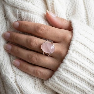 Könnte beinhalten: Ein Silberring mit einem großen, ovalen Rosenquarz-Edelstein. Der hellrosa Stein ist in einem schlichten Silberband gefasst. Der Ring wird an einer Hand gezeigt, mit einem weißen Strickpullover im Hintergrund.