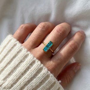 Pode incluir: Um anel de ouro com uma pedra retangular de turquesa. O anel tem uma banda fina e uma moldura de bisel. A turquesa tem uma cor azul-esverdeada vibrante com padrões de matriz natural. O anel é usado em um dedo.