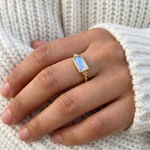 May include: A gold ring featuring a rectangular, iridescent gemstone. The ring has a delicate band and small decorative beads on the sides of the gemstone. The gemstone displays a range of blue and white hues. The ring is worn on a hand.