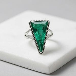 May include: A silver ring with a large, triangular emerald-green gemstone. The gemstone is framed by a beaded border. The ring has a split shank design, set against a white background.