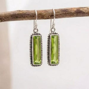 May include: A pair of silver earrings with rectangular, light green gemstones. The earrings are suspended from a silver hook and have a beaded border. The earrings are displayed on a brown branch against a white background.