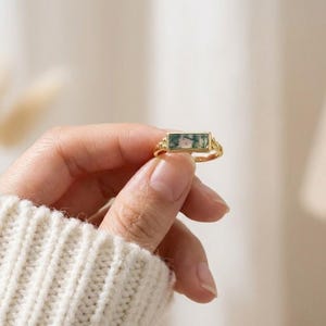 Puede incluir: Un anillo dorado con una piedra rectangular de ágata musgosa. La piedra presenta patrones verdes y blancos. El anillo tiene detalles decorativos en los laterales. El anillo lo sostiene una persona que lleva un suéter blanco.