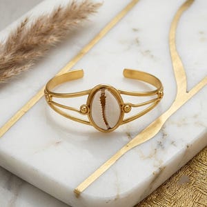Op de afbeelding: Een goudkleurige metalen manchetarmband met een centrale ovale zetting die een enkele witte kaurischelp bevat. De armband is gemaakt van twee dunne, parallelle draden die samenkomen bij de schelpzetting, geaccentueerd met kleine metalen bolletjes.
