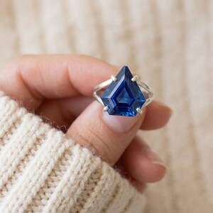 May include: A silver ring featuring a large, faceted, deep blue gemstone. The gemstone is cut into a unique geometric shape, with a pointed top and multiple facets. The ring is held against a soft, cream-colored background.