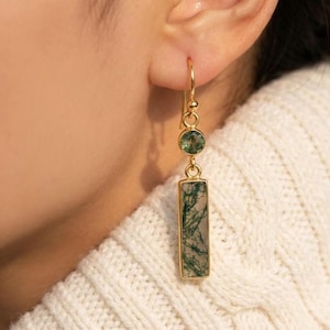 May include: Gold-toned dangle earrings featuring a round green gemstone and a rectangular moss agate stone. The earrings are suspended from a gold hoop and are worn on the ear. The moss agate has a natural, organic pattern.