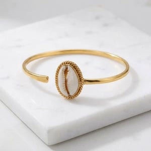Op de afbeelding: Een goudkleurige metalen armband met een centraal ovaal kauri-schelp element. De schelp is wit met een natuurlijke bruine spleet in het midden en is omlijst door een gedraaide goudkleurige metalen rand. De armband heeft een gladde, open band.