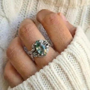 Può includere: Un anello d'argento con una pietra preziosa di agata muschiata verde a forma di pera. L'anello presenta dettagli floreali e di foglie intorno alla pietra, creando un design ispirato alla natura. L'anello è indossato su un dito.