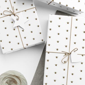 May include: White gift boxes with a repeating bee pattern, tied with twine and a tag. The boxes are presented on a white surface. A ball of beige yarn is in the lower left corner.