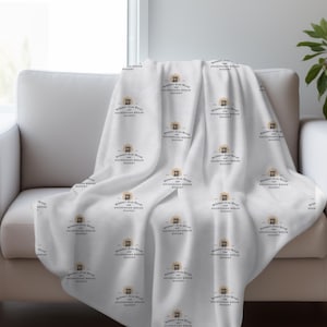 Pode incluir: Cobertor de lã branca com um padrão repetido de um logotipo de padaria. O logotipo apresenta um edifício com uma placa que diz "Sweet Home Bakery".