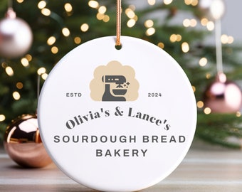 Puede incluir: Adorno navideño de cerámica blanca con un logotipo marrón y blanco que dice "Olivia's & Lance's Sourdough Bread Bakery" y "ESTD 2024". El logotipo también incluye una imagen estilizada de una batidora de pie.