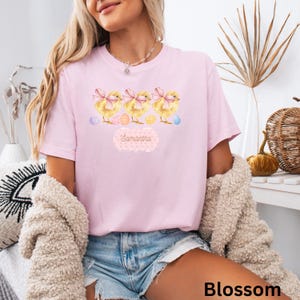 以下が含まれることがあります： ピンクのTシャツに、ピンクのリボンをつけてイースターエッグを持った黄色いひよこ3羽のグラフィックがプリントされています。Tシャツには「Samantha」という文字がプリントされています。