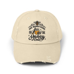 Peut inclure: Une casquette de baseball beige usée avec un motif d'abeille noir et jaune et le texte "She works hard for the honey".