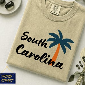 Peut inclure: Un t-shirt beige à manches courtes avec un col rond et une étiquette de marque Comfort Colors. L'avant présente le texte noir South Carolina en écriture cursive à côté d'un palmier graphique aux palmes bleu foncé et au tronc orange.