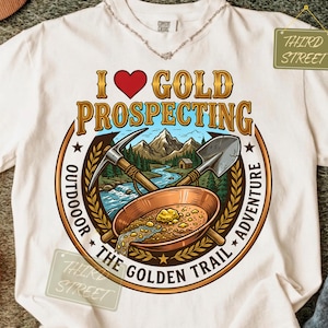 Peut inclure: Un t-shirt couleur crème avec une illustration représentant une batée, une pioche et une pelle. Le texte indique I HEART GOLD PROSPECTING, OUTDOOR THE GOLDEN TRAIL ADVENTURE et THIRD STREET. Le dessin montre des montagnes, une rivière et une cabane.