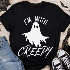 Peut inclure: Un t-shirt noir à manches courtes avec un motif blanc représentant un fantôme au centre. Au-dessus du fantôme se trouve le texte "I'M WITH" et en dessous le texte "CREEPY" dans une police blanche stylisée. Le t-shirt a un col rond et des manches retroussées.