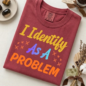 Könnte beinhalten: Ein kastanienbraunes Kurzarm-T-Shirt mit Rundhalsausschnitt. Auf der Vorderseite befindet sich der gelbe, violette, hellblaue und orangefarbene Schriftzug "I Identify As A PROBLEM", der von kleinen orangefarbenen "x"-Symbolen umgeben ist.