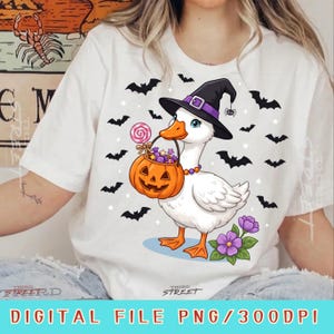Peut inclure: Un t-shirt blanc avec un motif représentant un canard blanc portant un chapeau de sorcière noir à bande violette. Le canard tient un seau citrouille orange rempli de bonbons violets et une sucette rose, entouré de chauves-souris et fleurs.