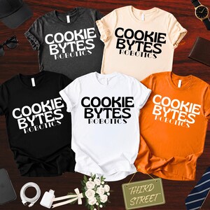 May include: Five short-sleeved t-shirts in charcoal, cream, black, white, and orange. Each shirt features the text "COOKIE BYTES ROBOTICS" printed in a bold, stylized font. The shirts are arranged on a dark wood surface with various accessories.
