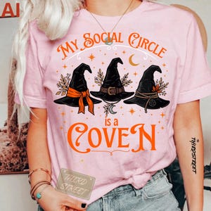 Peut inclure: Un t-shirt rose clair avec un motif représentant trois chapeaux de sorcière noirs aux accents orange et marron. Le texte "MY SOCIAL CIRCLE is a COVEN" est écrit en orange, entouré de petites étoiles et d'un croissant de lune.