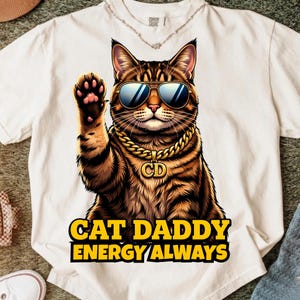 Peut inclure: Un t-shirt couleur crème avec un motif de chat tigré brun portant des lunettes de soleil teintées de bleu et une chaîne dorée avec un pendentif CD. Le chat agite une patte. Sous le chat, le texte CAT DADDY ENERGY ALWAYS est écrit en jaune.
