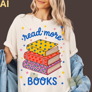 Peut inclure: Un t-shirt crème avec un motif de trois livres empilés aux motifs d'étoiles bleues, de cœurs rouges et d'imprimé léopard rose. Le texte "read more" est écrit en bleu au-dessus des livres et "BOOKS" en bleu en dessous, entouré de petites étoiles.