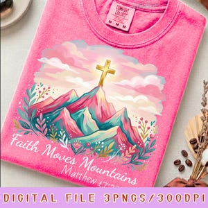 Peut inclure: Un t-shirt rose vif avec un motif représentant une croix dorée au sommet de montagnes stylisées sous un ciel nuageux rose et blanc. Le design inclut des accents floraux colorés et le texte blanc "Faith Moves Mountains Matthew 17:20".