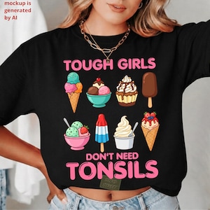 May include: A black t-shirt featuring a graphic print of various ice cream treats and desserts. The text reads TOUGH GIRLS DON'T NEED TONSILS in pink capital letters. The design includes ice cream cones, sundaes, a popsicle, and a cupcake.