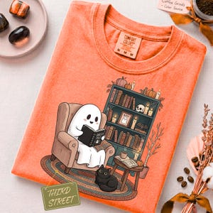 Peut inclure: Un t-shirt orange avec une illustration d'un fantôme blanc assis dans un fauteuil marron lisant un livre intitulé Ghostly Tales. À côté du fauteuil se trouve une bibliothèque remplie de livres, bougies, un crâne et un chat noir sur un tapis.