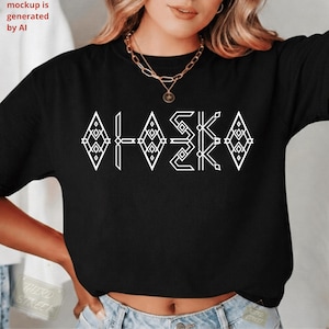 May include: A black short-sleeved t-shirt featuring the word ALASKA printed in a white geometric, diamond-patterned font across the chest. The shirt is paired with a gold-toned layered chain necklace with a small circular pendant.