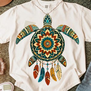 Peut inclure: Un t-shirt à manches courtes de couleur crème arborant un grand motif de tortue marine. La carapace de la tortue présente un motif mandala coloré dans des tons sarcelle, orange et jaune. Cinq plumes pendantes rouges, sarcelle et jaunes.