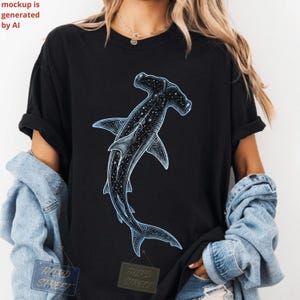 Peut inclure: Un t-shirt noir à manches courtes orné d'un grand motif de requin-marteau. Le requin est illustré avec un motif céleste sombre rempli d'étoiles et de constellations blanches, souligné par une lueur bleu clair sur le tissu noir.
