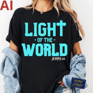 Peut inclure: Un t-shirt noir à manches courtes avec le texte cyan vif LIGHT OF THE WORLD, une croix intégrée dans la lettre T et la référence JOHN 8:12 imprimée en blanc en dessous. Le t-shirt est porté avec une veste en jean bleu clair.