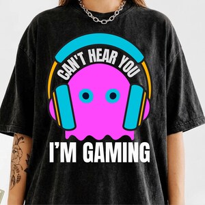 May include: A black acid-wash t-shirt featuring a graphic of a bright pink ghost wearing large blue and orange headphones. The text "CAN'T HEAR YOU" is arched above the ghost, and "I'M GAMING" is printed in white capital letters below the graphic.