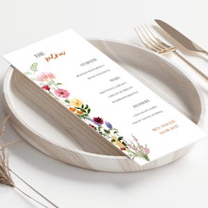Może przedstawiać: Karta menu weselnego z kwiatowymi akcentami, z napisem "menu" w kursywie. Menu zawiera przystawki, dania główne i desery. Karta znajduje się na drewnianym talerzu z widelcem i nożem.