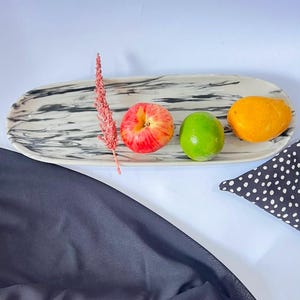 May include: An elongated oval serving tray with a white and black marbled pattern. The tray holds a single pink and yellow apple, a bright green lime, a yellow mango, and a tall, thin, textured pink dried floral stem placed horizontally.