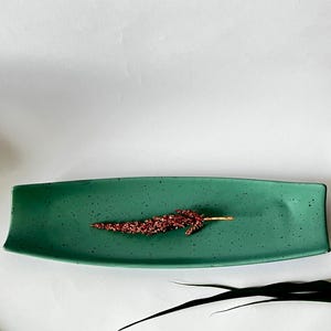 Op de afbeelding: Een rechthoekig groen keramieken dienblad met licht gebogen randen en een gespikkelde afwerking. In het midden van het dienblad ligt een enkele, slanke, roodbruine decoratieve stengel bedekt met fijne, getextureerde deeltjes, die doet denken aan een gedroogd botanisch element.