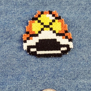Peut inclure: Un patch en forme de champignon pixelisé avec un motif noir, blanc, orange et jaune. Le patch est sur un fond en jean bleu.