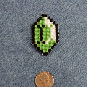May include: Green and white pixelated gem with a black outline, resembling a video game item. The gem is on a blue denim background.