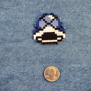 Peut inclure: Un patch en forme de champignon pixelisé bleu et blanc avec des détails noirs. Le patch est sur un fond en jean bleu.