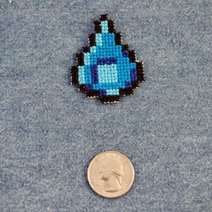 May include: A blue and black pixelated diamond-shaped patch, reminiscent of a video game item, is displayed on a denim background.