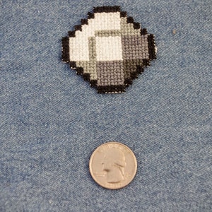 Peut inclure: Un petit motif de point de croix en forme de diamant, gris et blanc, sur un fond de denim bleu. Une pièce de monnaie américaine de 25 cents est placée sous le motif pour l'échelle.