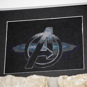 以下が含まれることがあります： アベンジャーズのロゴが刺繍された黒と白の額装された画像。ロゴはスタイリッシュな「A」で、周りを円が囲んでいます。円は青と白のパターンで満たされています。