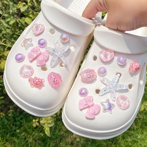 May include: White clogs decorated with pink and iridescent charms. The charms include starfish, hearts, bows, and other sea-themed embellishments. The clogs are shown outdoors on a bed of green grass.